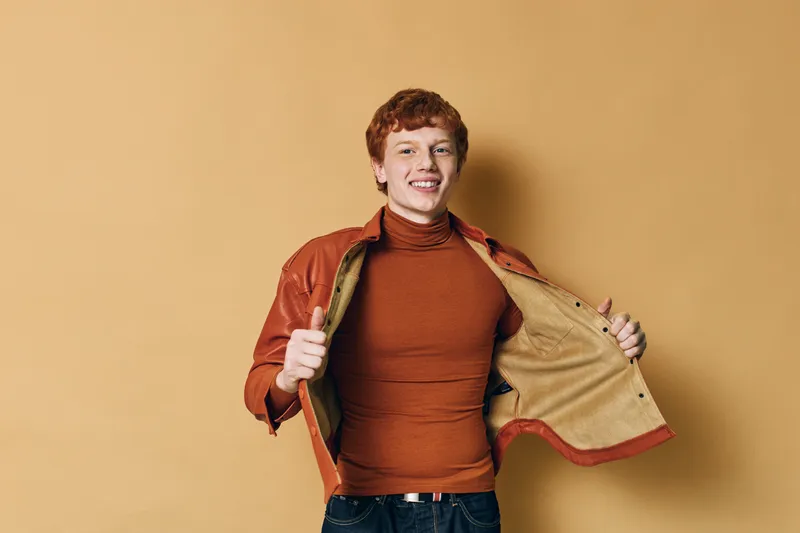Young man with red hair smiling while wearing brown turtleneck and leather jacket on beige background. Casual style, confident pose, isolated portrait with positive expression.