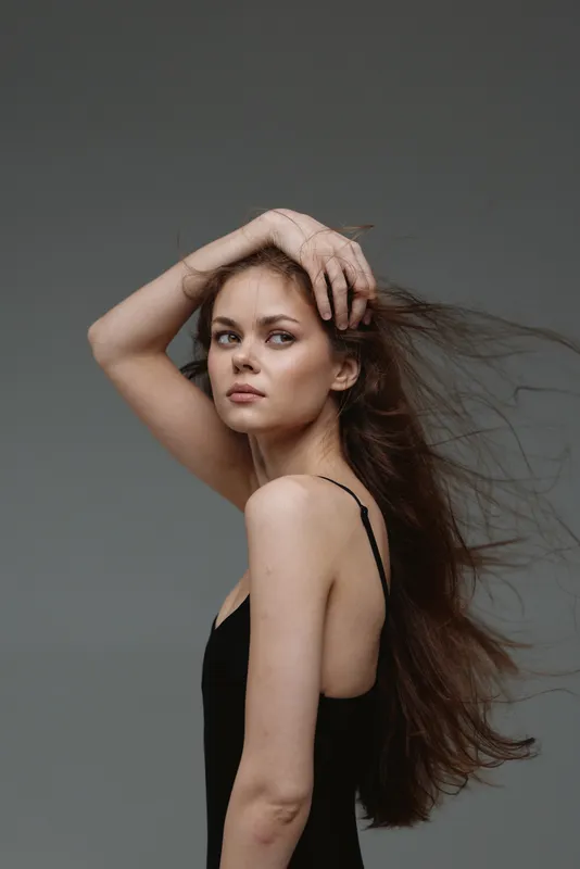 Portrait of a young woman with long flowing hair and a black sleeveless dress posing against a gray background