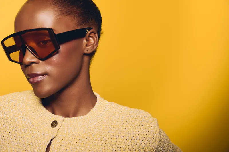 Portrait of a confident dark skinned African woman wearing large sunglasses and a yellow knitted sweater against a bright yellow background. Concept of fashion and style.