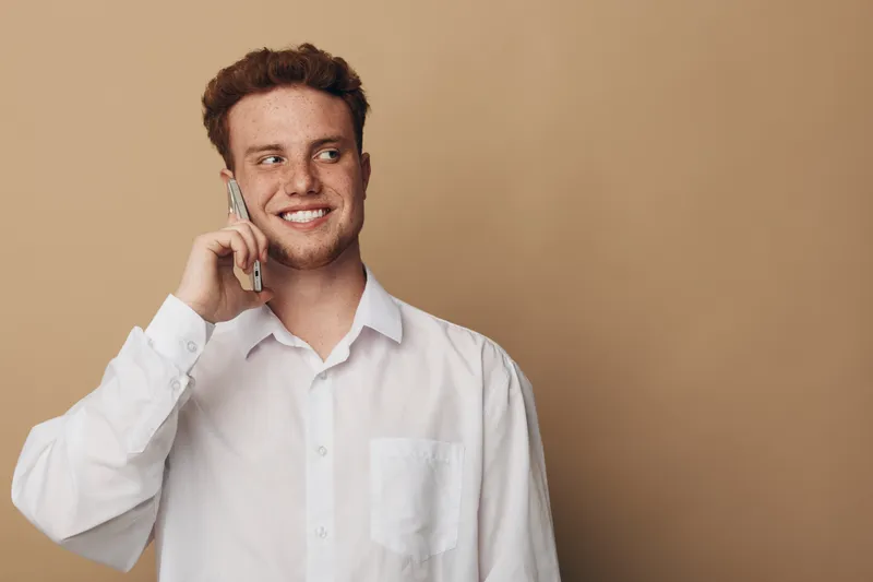 Smiling young man with red hair wearing white shirt talking on smartphone against beige background. Concept of communication, technology, casual lifestyle, and positive emotions.