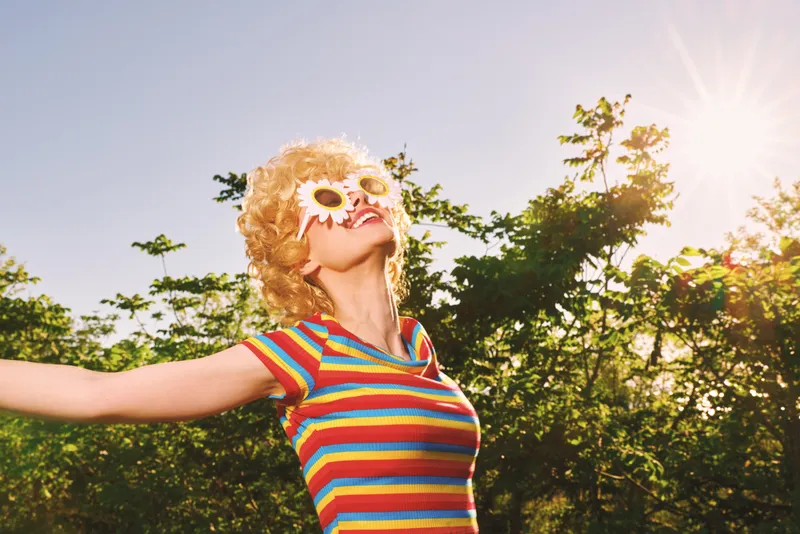 A joyful woman outdoors wears a striped shirt and holds a flower over her eye, basking in bright sun. A playful, retro vibe with vibrant colors. The scene evokes a carefree lifestyle and bold style