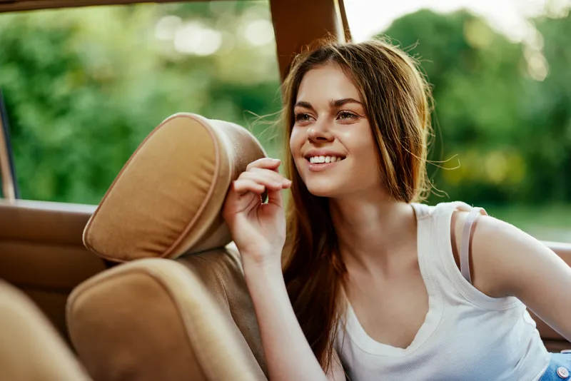 Young woman smiling in car, enjoying summer outdoors, with green background and warm sunlight, representing joy, freedom, and adventure in casual attire.