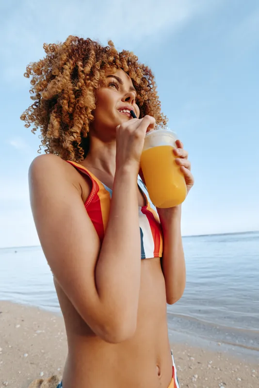Woman with curly hair on beach, ocean horizon behind, sipping juice drink, relaxed smile, warm sunlight, summer vacation mood, natural skin tones, bright sky
