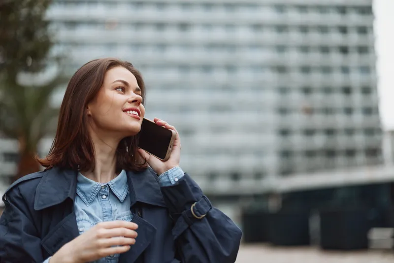 Young woman with brown hair wearing a blue coat talks on a smartphone outdoors in an urban environment with a blurred building background.