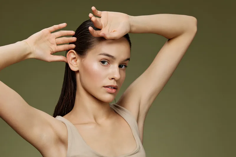 Young woman with a smooth complexion posing gracefully against a solid green background, showcasing a minimalistic fashion style and a subtle expression of confidence
