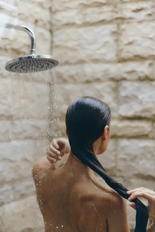 woman showering outdoors with wet hair under rain showerhead, natural stone wall background, skincare and wellness concept, serene mood
