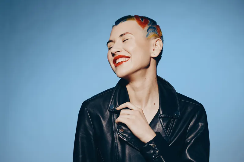 Smiling woman with colorful short hair and bright red lipstick wears black leather jacket against plain blue background, expressing joy and unique style in modern portrait.
