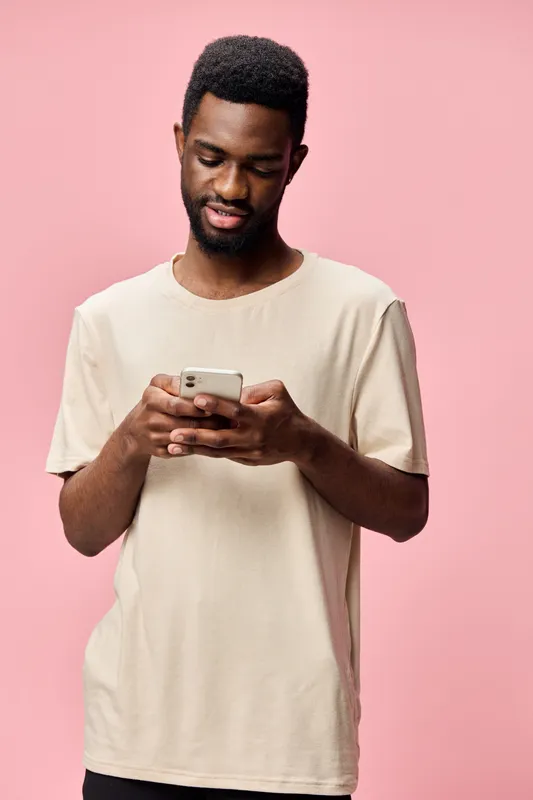 Young Black man in casual beige shirt using smartphone against pink background, conveying relaxed and modern lifestyle, technology and social media engagement