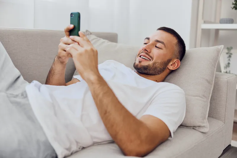 Relaxed man enjoying his smartphone while lying on a cozy couch, embodying modern lifestyle and leisure trends in 2025, embracing comfort and technology.