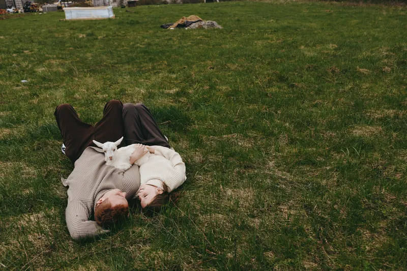Lifestyle scene of a person and a playful goat kid resting on green grass, outdoor calm moment, affection and connection, simple countryside joy, relaxed cuddle with animal companion.