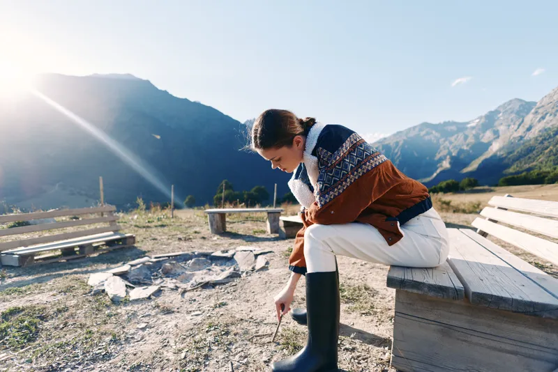 Woman in a warm sweater and boots sitting on a wooden bench in the mountains, enjoying outdoor nature in autumn sunlight near a fire pit for travel, rest and relaxation.