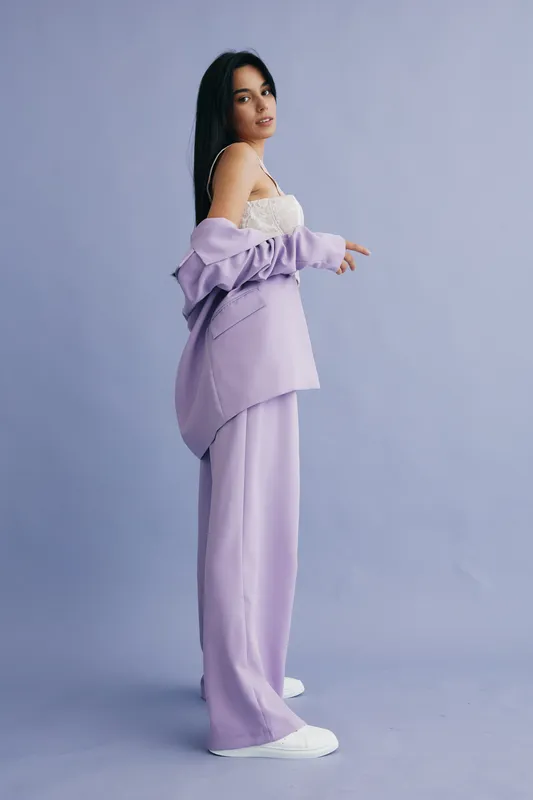 Stylish woman in lavender suit and white lace top poses confidently against soft purple background, showcasing contemporary fashion and casual elegance indoors.