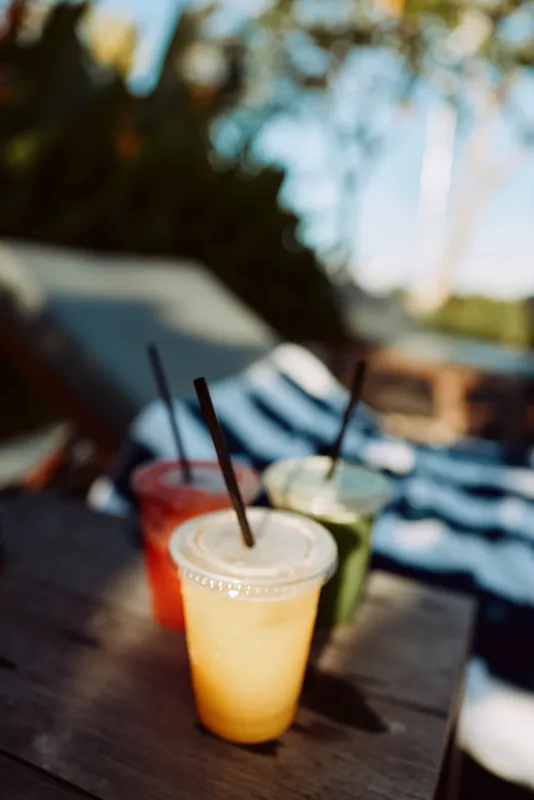 Three colorful fresh juice drinks in plastic cups with straws on wooden table outdoors. Healthy lifestyle concept with natural light and blurred background for summer relaxation.