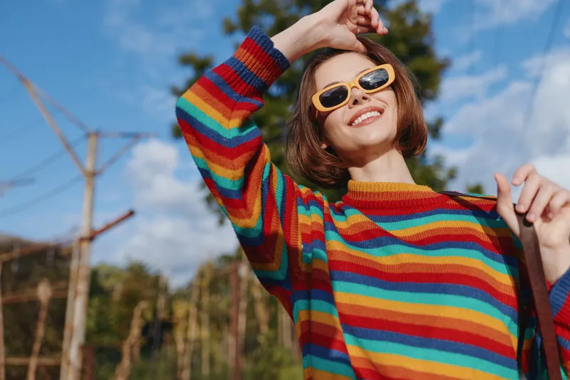 Young woman in her twenties with short hair wearing a rainbow sweater and yellow sunglasses, smiling and leaning back outdoors on a sunny day, casual outfit, lifestyle fashion portrait.