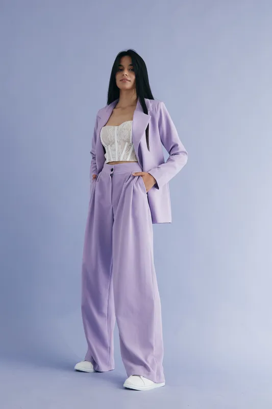 woman in lilac suit standing confidently against a pastel background wearing a white lace top and white sneakers, showcasing modern fashion style and casual elegance