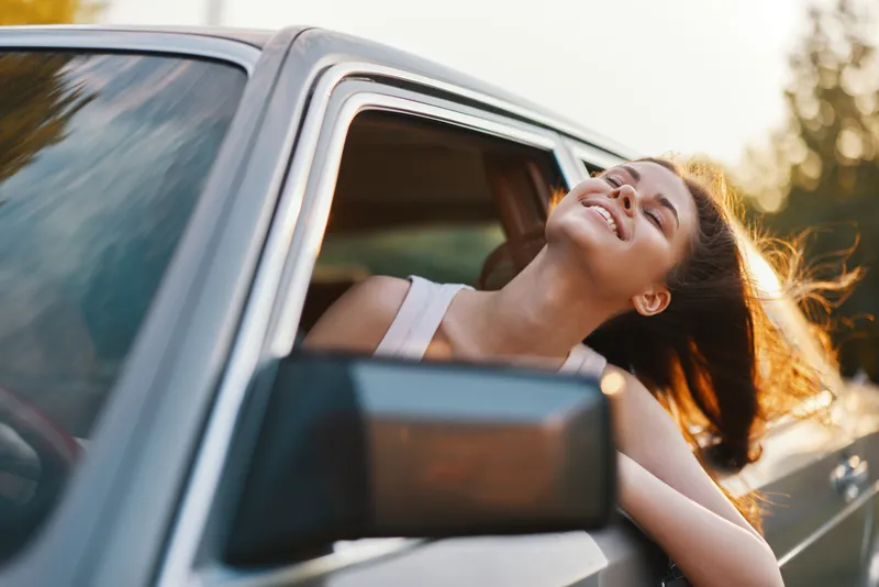 Young woman enjoying a carefree drive with her hair flowing in the wind, embodying freedom and joy, with a scenic background in warm sunset colors.
