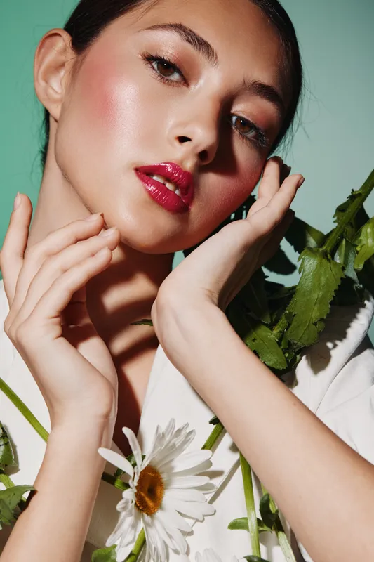 Close-up of a young woman with glossy red lips and dewy skin posing with white daisies against soft green background, embodying creative beauty and fashion concepts.