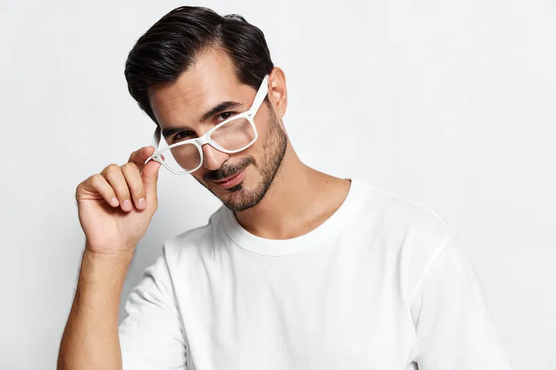 Stylish man with glasses poses confidently, adjusting eyewear while looking playfully at the camera, dressed in a casual white t-shirt against a light background.