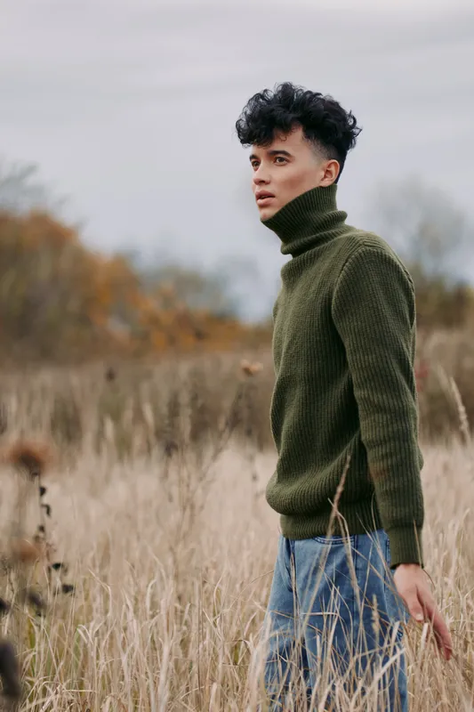 A young man in a textured olive turtleneck stands in tall dry grass, gazing thoughtfully into the distance, an authentic, grounded portrait captured with soft natural light and honest mood.
