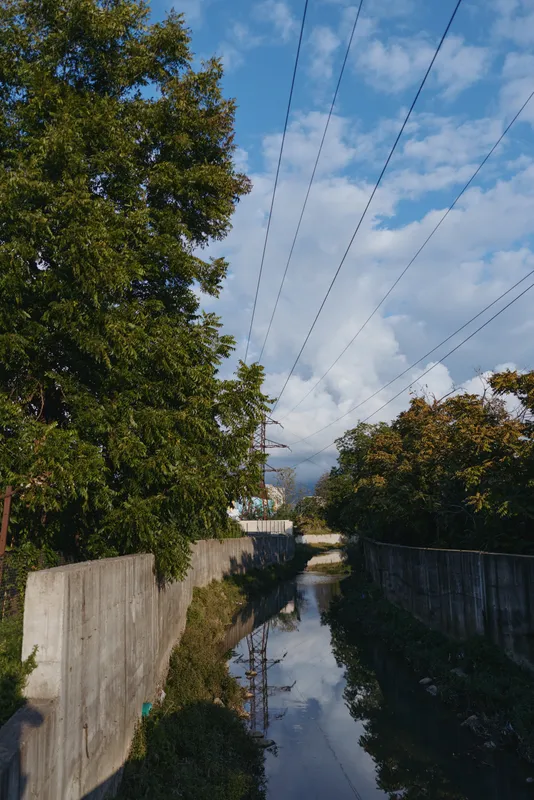 Concrete canal, storm clouds, overhead power lines, water reflection and riverside trees in an urban drainage channel. Empty suburban scene with moody sky for infrastructure or environment study.
