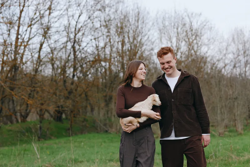 Cheerful couple outdoors, smiling with goat kid, casual outfits, sunlit meadow, gentle breeze, candid lifestyle moment, natural colors, surrounding trees, peaceful countryside scene