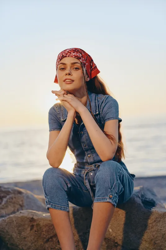 young woman wearing red bandana and denim jumpsuit sitting on rocks near calm sea during golden hour with relaxed expression and natural light