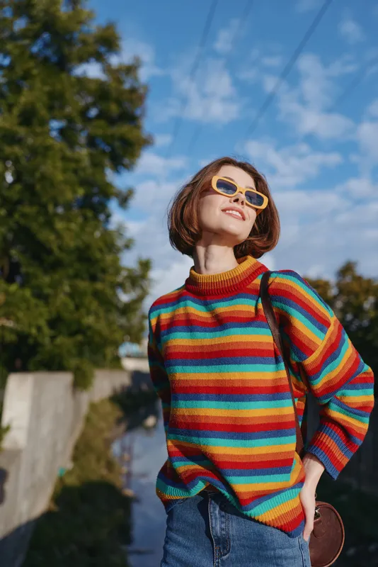 Young woman in a colorful striped sweater and sunglasses smiling with bob haircut, casual outfit and denim jeans on a sunny day outdoor portrait along a suburban walkway, travel lifestyle.