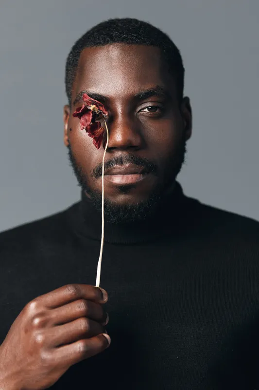 Creative conceptual portrait of African man holding dried flower in front of face with calm expression, dark skin and black turtleneck against neutral background in studio.