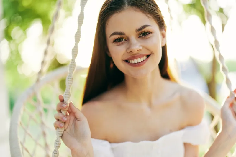 Happy young woman sitting on a swing, wearing a white off the shoulder dress, smiling in a sunny outdoor setting with a blurred green background
