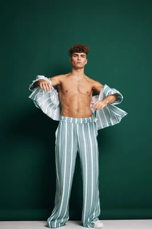 Young shirtless man posing in striped loose pants and matching open shirt isolated green background. Fashion confident urban style model with curly hair and barefoot in studio.
