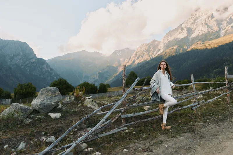 Woman in a light sweater standing beside a rustic wooden fence in a mountain landscape. Outdoor lifestyle photo highlighting nature, travel, and calm mood with soft daylight.