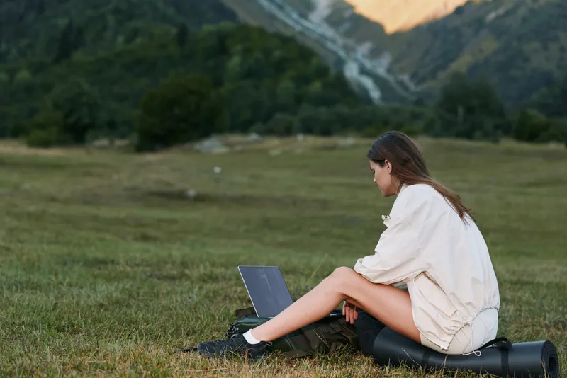 A focused young woman sits outdoors on a grassy field with a laptop, closely engaged in work and enjoying a calm, productive moment in nature, away from a busy indoor environment.