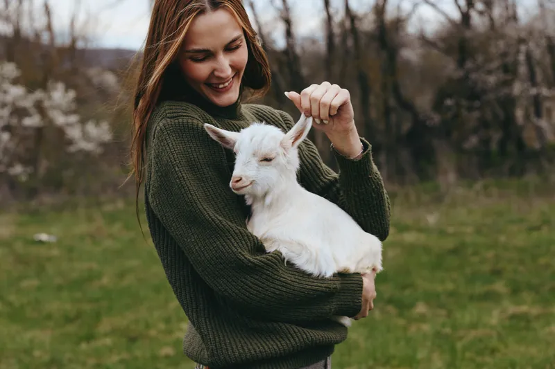 Woman smiling outdoors while holding a white goat kid in a grassy field. Lifestyle scene emphasizes gentle bonding with animals, nature, and wellness. Soft natural light highlights warmth, care, and