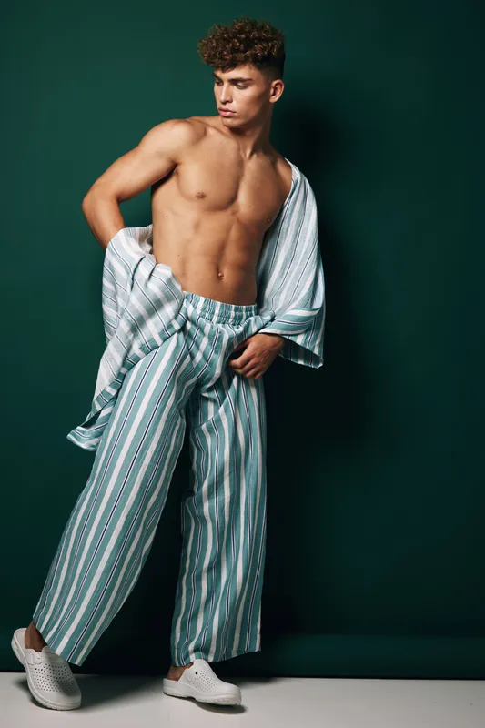 Young man with muscular body posing in striped loose pants and textile shirt partially off shoulder on dark green background in the studio. Curly hair model in full length with white shoes.