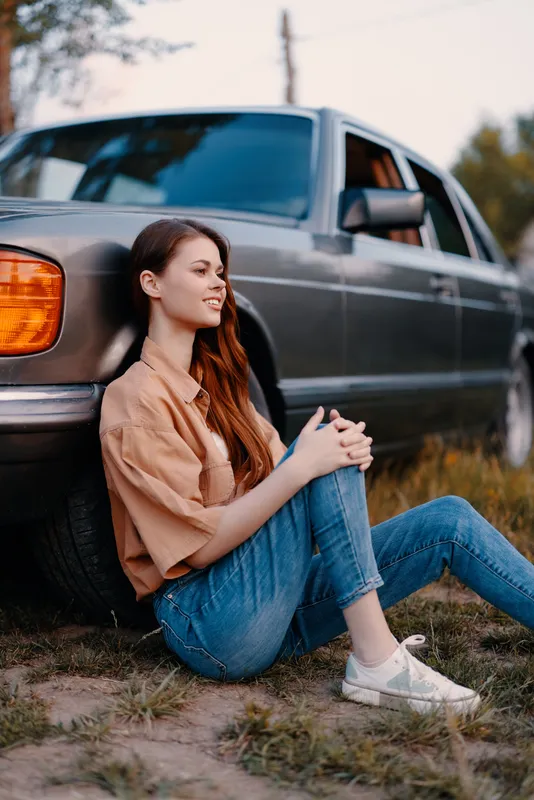 Young woman sitting by a vintage car in casual attire, showcasing a relaxed vibe and comfortable style against a natural background, perfect for lifestyle concepts.