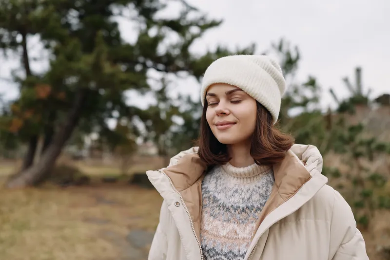 Woman outdoors in a winter coat and beanie, eyes closed in a serene moment. A peaceful portrait amid nature with soft light, calm mood, and fashionable yet cozy vibe.