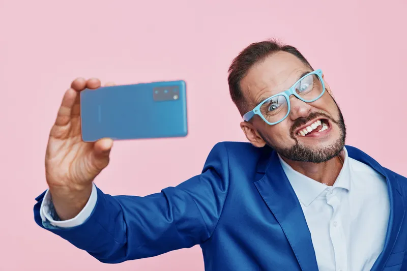 A professional business man takes a lively selfie against a solid pink backdrop, showcasing confidence, enthusiasm, and a modern, upbeat mood in a vivid studio setting.