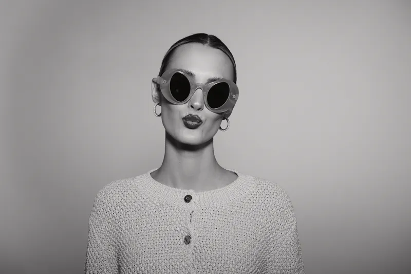 Portrait of a young Caucasian woman with fair skin wearing sunglasses and a knitted sweater, posing with puckered lips in a studio setting.
