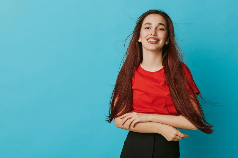Young woman with long hair and red shirt smiling confidently against blue background. Portrait of happy female with casual style and natural expression in studio setting.