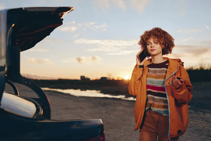 Woman lifestyle outdoors beside car at sunset on road, relaxed face, curly hair blowing, orange jacket, striped sweater, holding backpack strap, warm golden sky, seaside horizon, calm