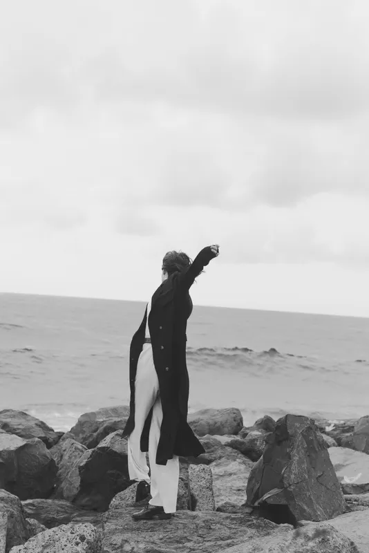 Couple embracing on a rocky shore by the sea, long coat and flowing dress in monochrome, capturing romance, wind, and tranquil coastal mood, and timeless atmosphere.