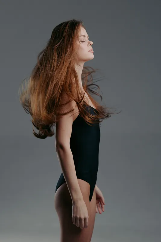 Young woman standing in black bodysuit with flowing hair and calm expression on gray background