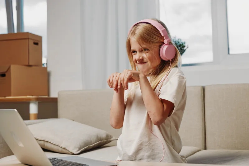 Young girl with pink headphones listens to music on laptop, dancing and smiling in cozy living room with cardboard boxes. Concept of childhood joy, home entertainment, and technology use.