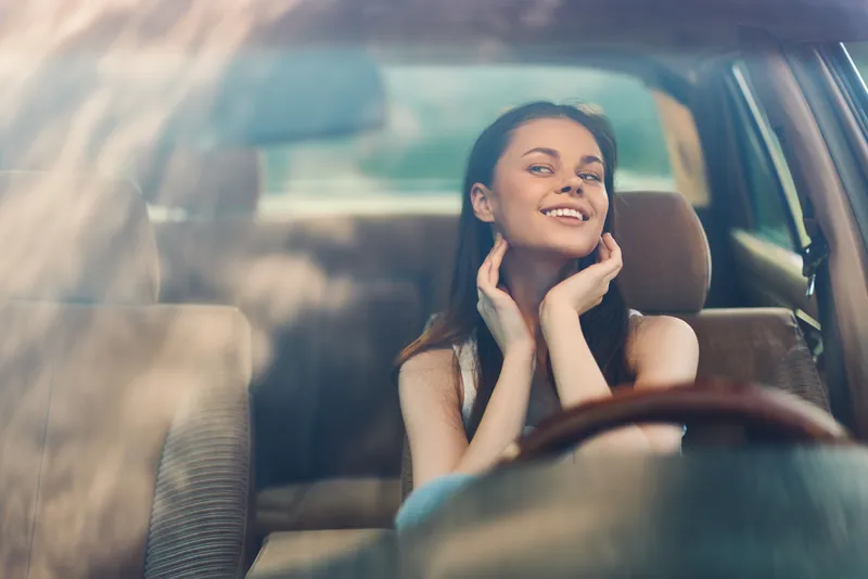 Young woman smiling in a car, enjoying the moment with a relaxed and happy expression in a sunny setting, emphasizing joy and freedom.
