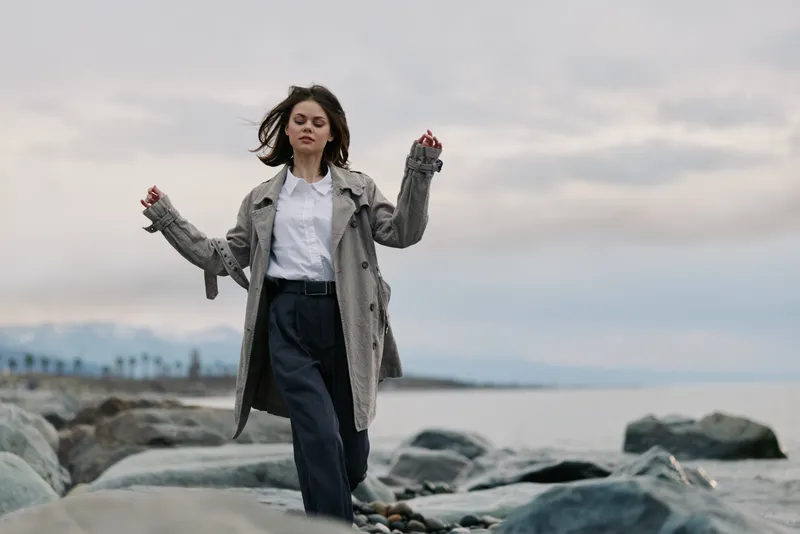 Young woman in casual clothes walks on rocky beach near calm sea under cloudy sky. Outdoor lifestyle, freedom, and nature connection concept with relaxed mood and natural environment.