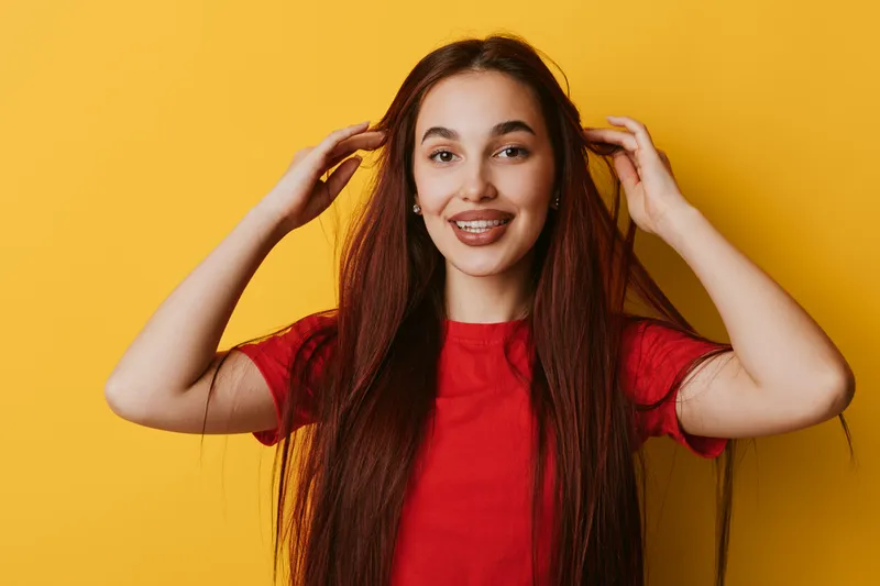 Young woman with long brown hair wearing red t shirt smiling and touching hair on yellow background. Portrait of happy female with natural makeup and casual style in studio.