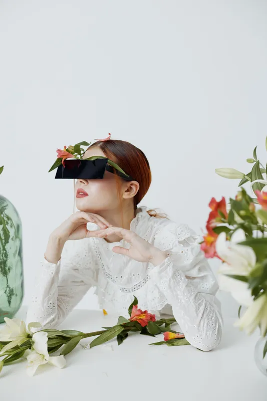 Young woman in floral attire with oversized sunglasses posing creatively among vibrant flowers in a minimalist setting, symbolizing spring s beauty and elegance