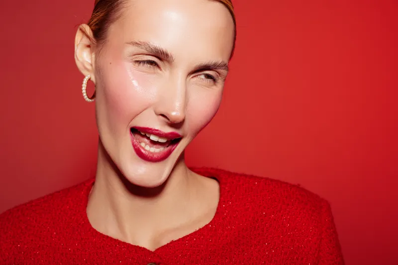 Close-up of a smiling woman with glossy skin and red lipstick wearing a red sweater and hoop earrings. Bright red background creates a vibrant and joyful mood. Beauty and fashion concept.
