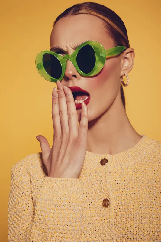 Surprised woman in green sunglasses covers mouth with hand, wearing yellow knitted sweater and gold hoop earrings. Studio portrait on yellow background with expressive facial emotion concept.