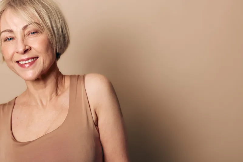 Smiling mature woman with short blonde hair wears beige tank top, standing against plain light brown background. Concept of aging beauty, confidence, and natural skin care.